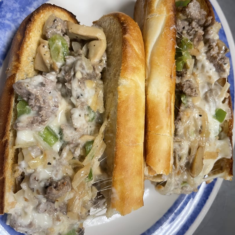 Two cheesy steak and mushroom subs with green peppers and onions on a blue and white striped plate.