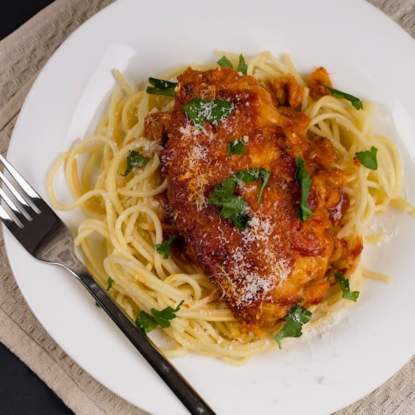 Chicken parmesan with spaghetti, red sauce, parsley, and grated cheese on a white plate.